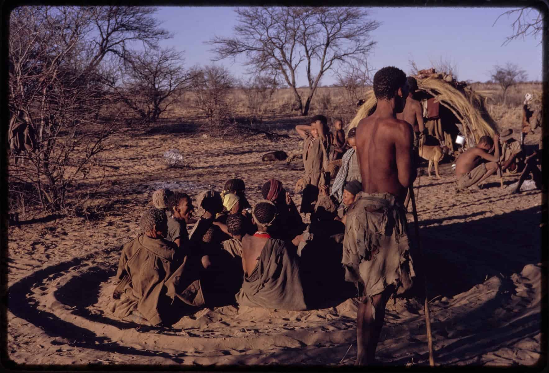 Group of women sitting inside curing dance circle. The deep sand furrows are made by the dancing healers. |Gaishay is standing nearby. 1961. Naron. Botswana/Ghanzi.